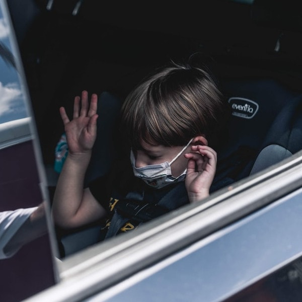 Durch eine heruntergelassene Scheibe sieht man einen kleinen Jungen mit OP-Maske im Kindersitz auf der Rückbank eines Autos.