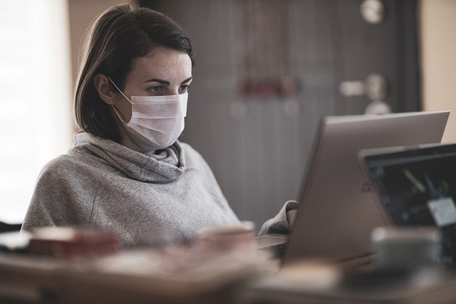 Zu sehen ist eine Frau mit einer OP-Maske, die vor einem Laptop sitzt.